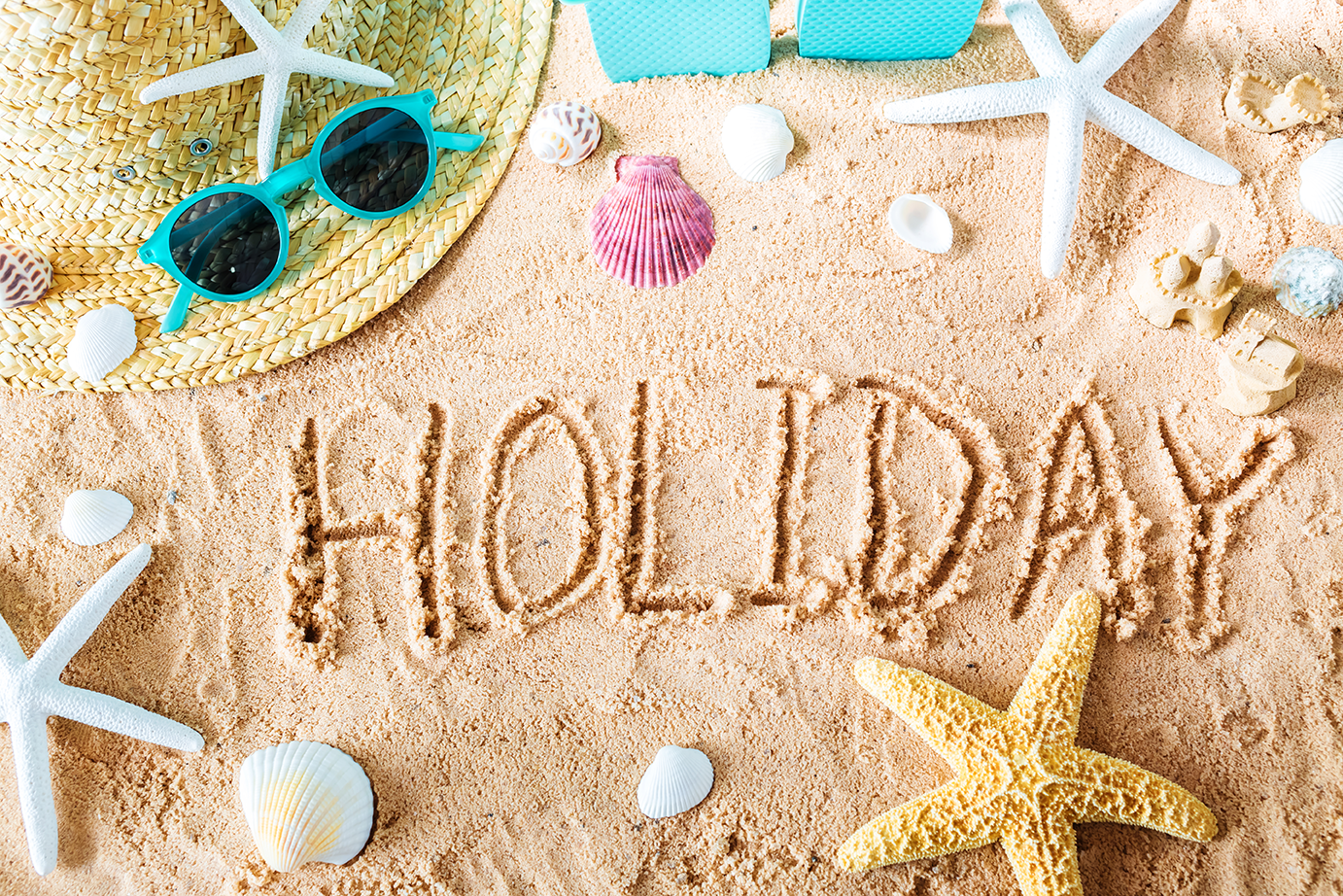 Beach and summer holiday lifestyle imagery — straw hat, sunglasses, starfish, and seashells arranged on sand with the word HOLIDAY written in it